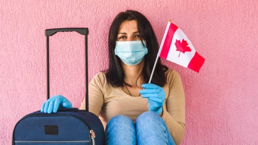 Woman sits in front of a pink wall holding her suitcase and a Canadian flag. She is wearing a face mask.
