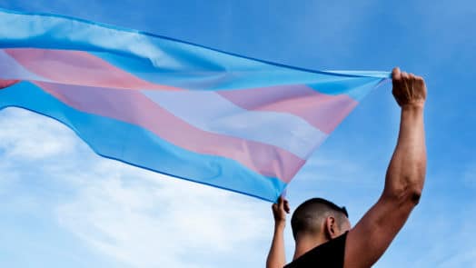 Man holding a trans pride flag against a blue sky