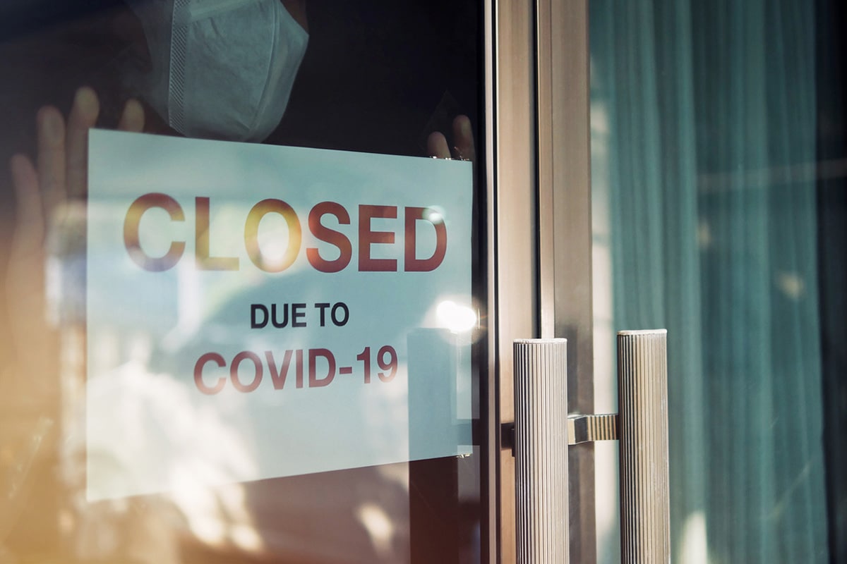 Person wearing surgical mask puts up "Closed due to COVID-19" sign on business door.