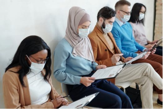 Multi-ethnic job candidates sitting in a row of chairs, wearing surgical masks.