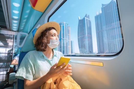 Woman in a protective medical mask rides the subway, holds a smartphone in her hands and looks out the window where the giant skyscrapers of Dubai rise.