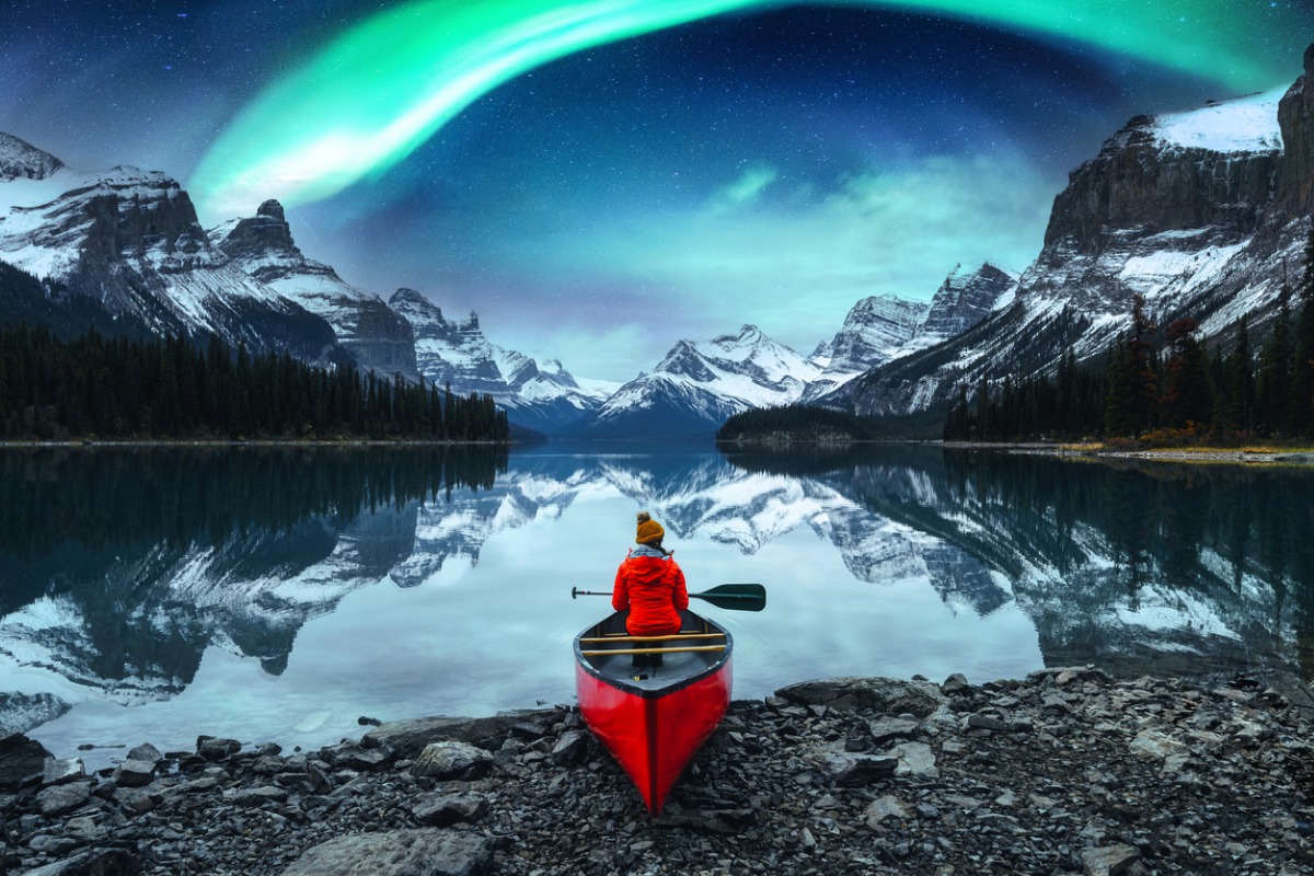 A person sits in a red canoe, which is banked on a rocky lakeshore in the mountains. The aurora borealis gleans overhead.