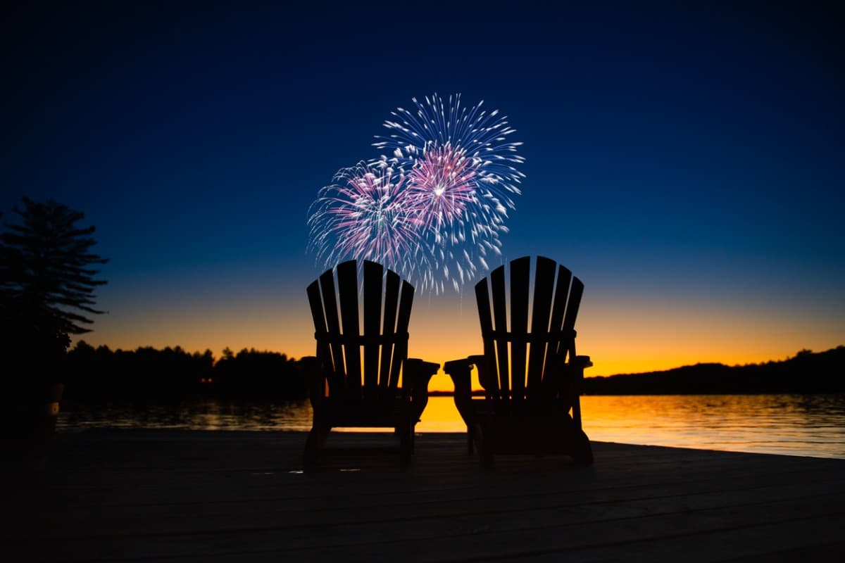 Two chairs overlooking a lake at sunset. Fireworks burst in the sky.