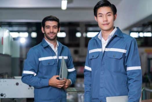 Two men in overalls in a factory, staring into the camera