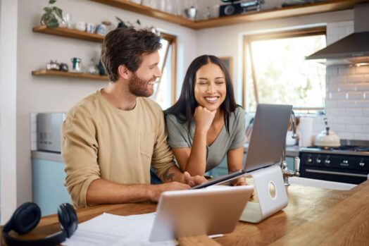 man and woman standing together smiling while woman looks at computer on table