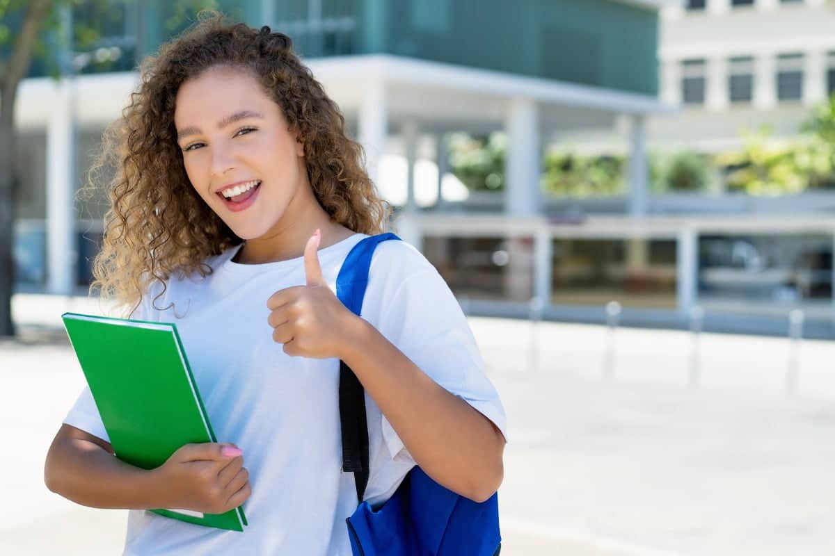 female student with backpack and paperwork holding one thumb up