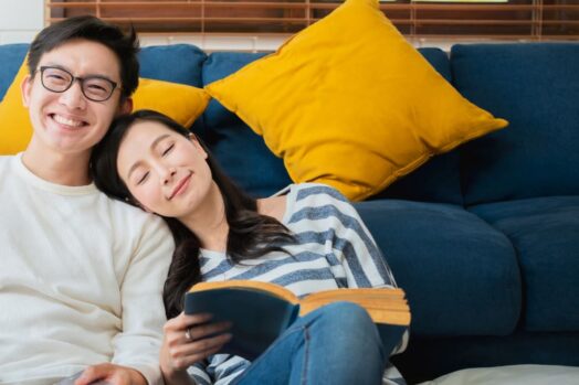 pareja sentada frente al sofá sonriendo.  Mujer leyendo libro y hombre sonriendo hacia adelante.