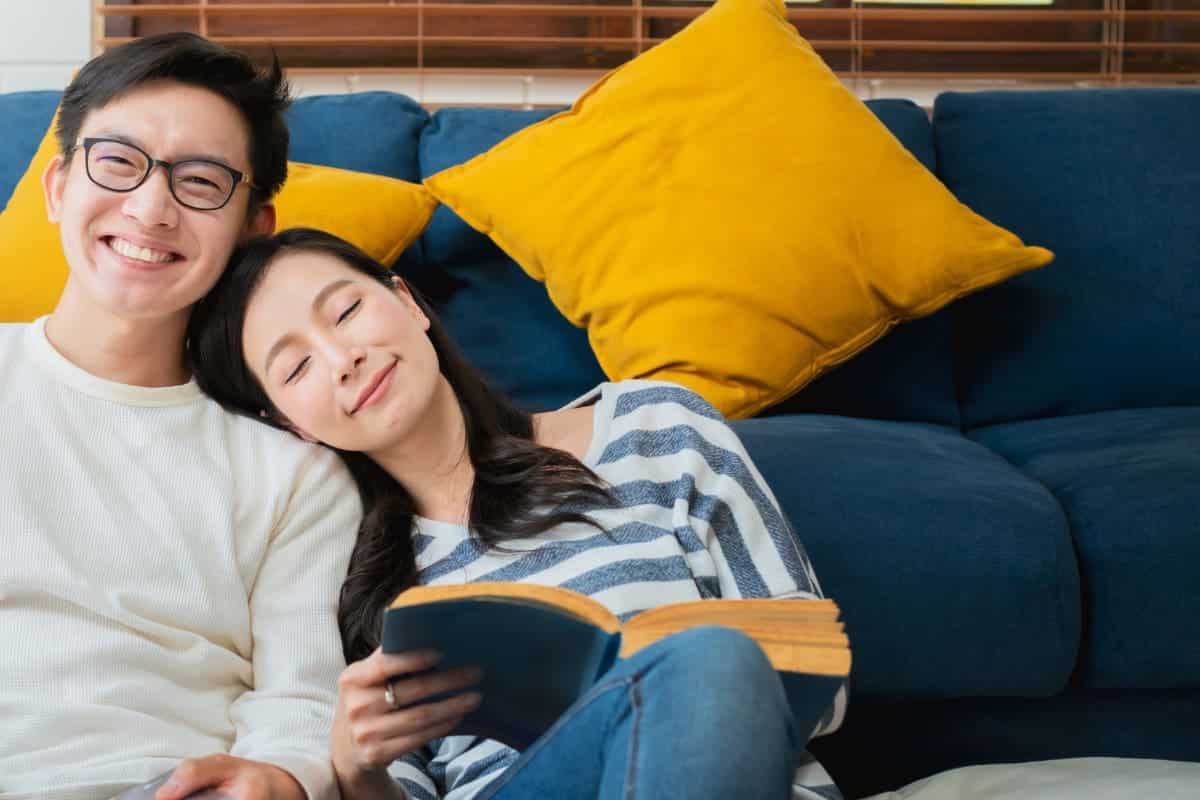 couple sitting in front of couch smiling. Woman reading book and man smiling forward.