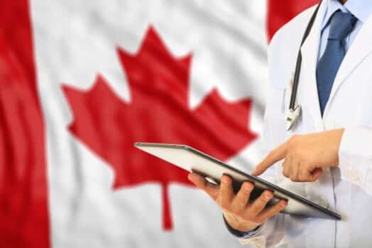 Doctor usando estetoscopio, ponting en un portapapeles.  Bandera de Canadá en segundo plano.