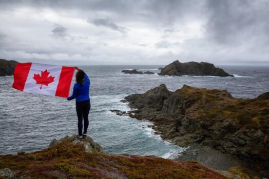 Person standing near body of water close to the coast of the ocean; waving a Canadian flag