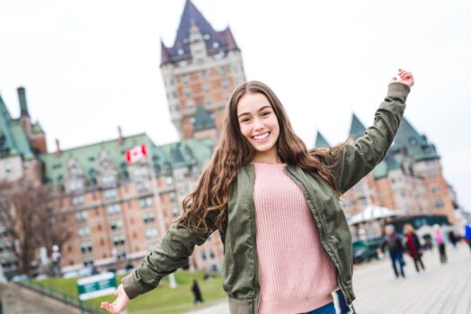 Estudiante de pie con los brazos extendidos y sonriendo frente a un edificio en Quebec.  Bandera canadiense en el fondo.