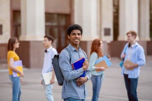 Un estudiante mira a la cámara mientras sus compañeros caminan detrás de él en un campus universitario.