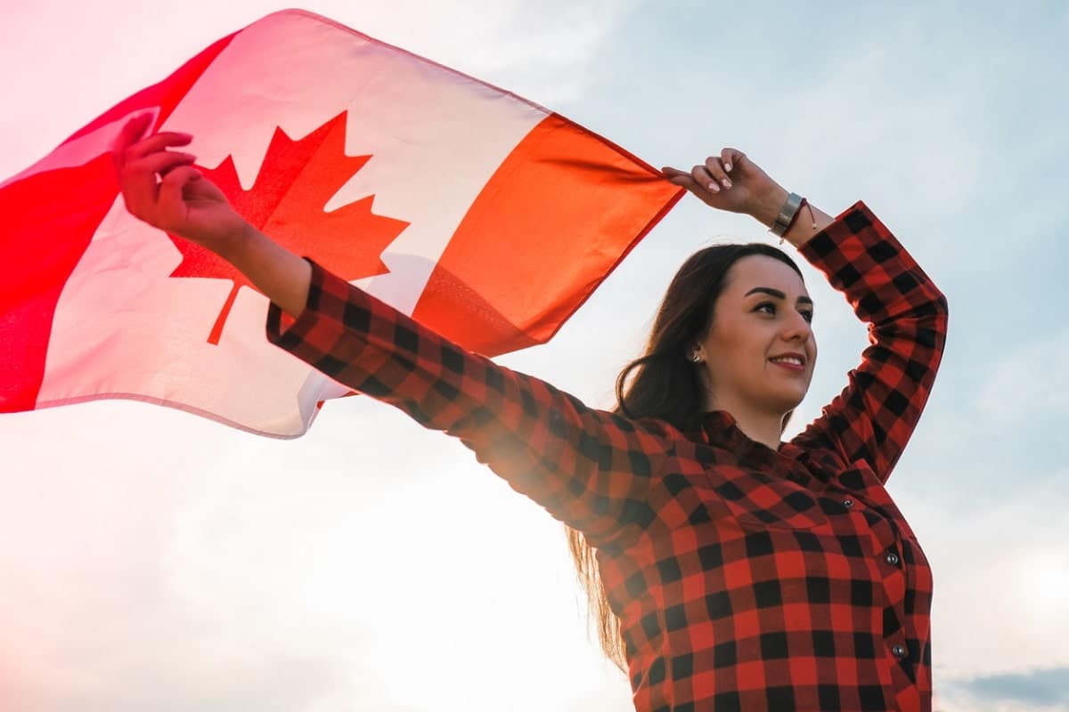 young millennial woman holding the Canadian flag behind her head as it waves in the wind.