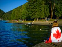 Patriot woman sitting on an old wooden pier over mazarine color water of the Cultus Lake and holding up a Canadian flag