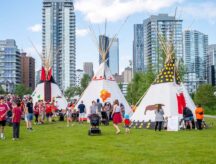 A group of people in a city park, in front of three indigenous teepees