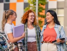 heerful female college students sitting in front of a university building, talking, smiling