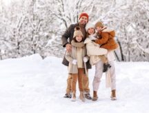 Outdoor portrait of family in warm clothes smiling at camera while spending time actively in winter snowy forest