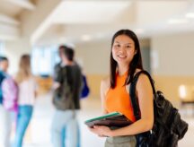 Portrait of a girl with a backpack and school supplies in the hallways of the university campus with her classmates in the background.
