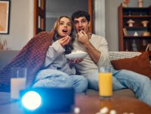 A young couple in love sitting on the sofa in the living room at home, eating popcorn, covered with a blanket and watching a movie on the projector.