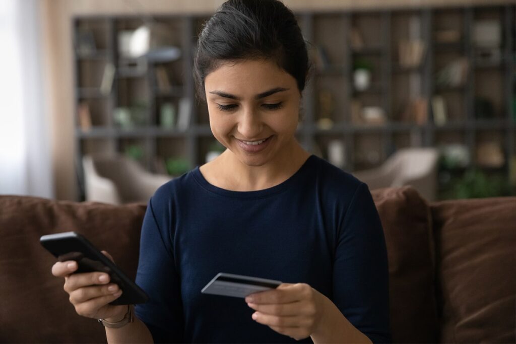 Woman sitting on couch looking at credit card in her left hand while holding a cell phone in her right hand