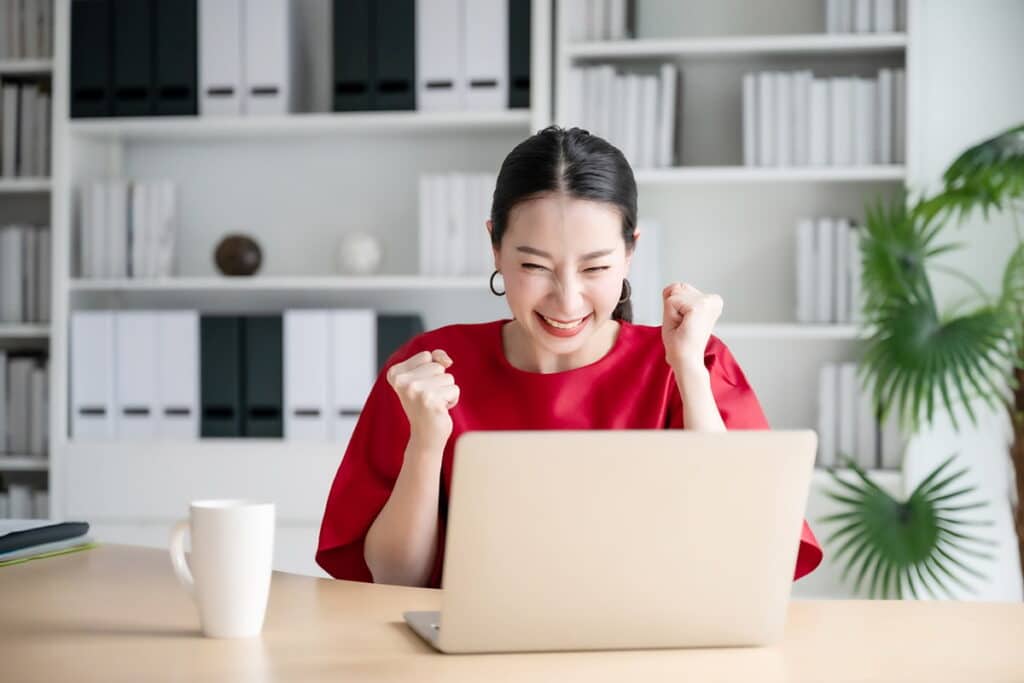 Mujer sentada en un escritorio mirando una computadora portátil y animando con entusiasmo, sonriendo y apretando ambas manos en puños