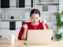 Woman sitting at desk looking at laptop and cheering in excitement, smiling and clenching both hands into fists