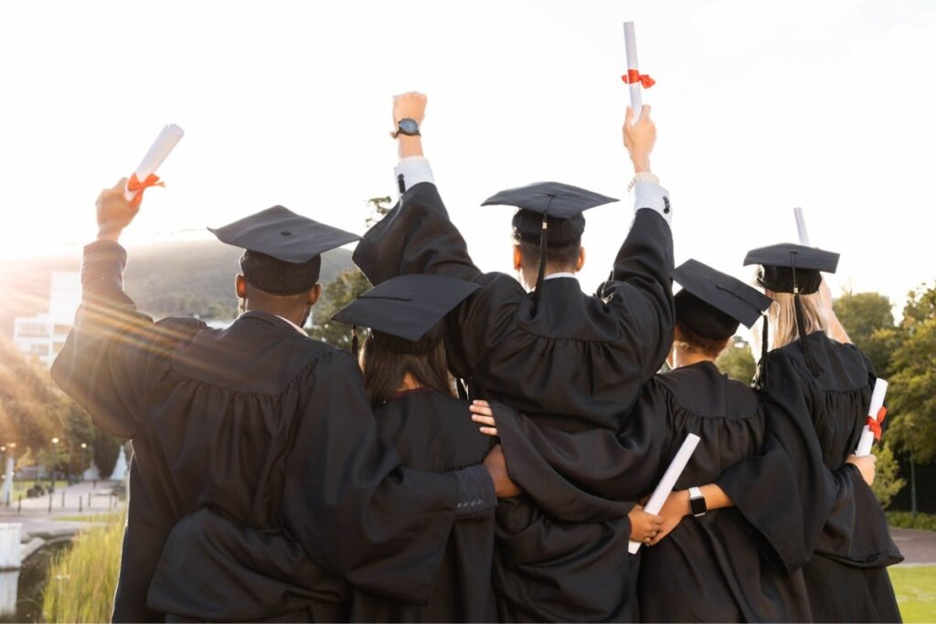 View from behind a group of five students celebrating their graduation while holding diplomas and wearing their caps and gowns.