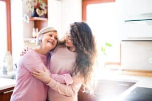 A young woman with long curly hair hugging an older lady with short, grey hair. They are standing in a kitchen, and Both are wearing pink