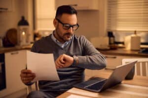 A man with glasses sitting at a table with a laptop, holding a piece of paper and checking the time on his watch.