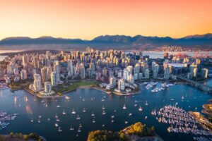 An aerial view of the downtown Vancouver skyline at sunset, with mountains visible in the background and sailboats in the foreground.