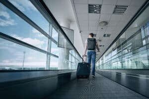 A man with a backpack and a luggage in hand at an airport terminal using a moving walkway.