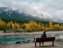 A woman sitting on a bench facing a river lined with yellow trees and mountains visible in the distance in Canmore, Alberta.
