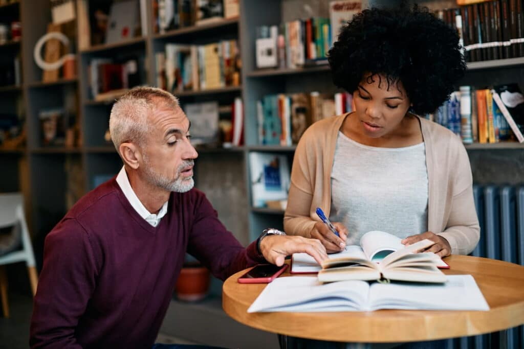 Older male tutoring a younger woman at a small round table covered with open books.