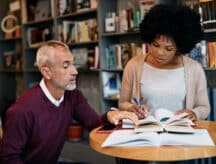 Older male tutoring a younger woman at a small round table covered with open books.