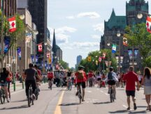 People both walking and riding their bikes on the street, surrounding by Canadian flags, near Parliament Hill.