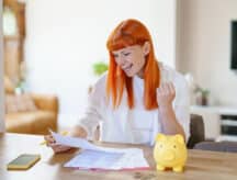 Women with orange hair sitting at a table, reviewing papers and ecstatically raising her fist. A yellow piggy bank sits next to her.