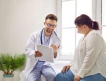 A smiling male doctor talking to a female patient as he motions toward the clipboard in his hand.