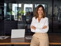 Smiling female office worker confidently leaning against a table with her arms crossed.