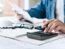A closeup shot of a person sitting at a table with their laptop and using a calculator, while surrounded by various papers.