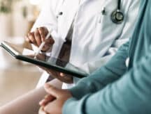 A close up shot of a doctor sitting with a patient, while holding and gesturing towards a tablet.