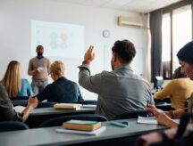 A group of students sitting in a classroom, with one raising his hand to answer a question.