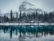 A shot of a wooden lodge nestled within snow-covered pine trees, with a still lake reflecting the trees and surrounding mountains.
