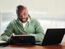 Smiling man sitting at a test, with a laptop to his side and a tablet in his hands.