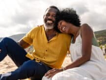 Middle-aged man and woman sitting together in the sand, laughing.
