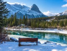 A sunny winter day at the river, with a bench in the foreground and a majestic mountain range in the background.