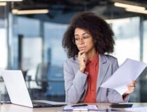 Woman sitting at a desk, looking quizzically at her laptop, while holding a piece of paper in her hand.