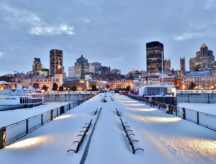 Downtown Montreal in the winter after sunset; the waterfront walkway and its benches are covered in snow.