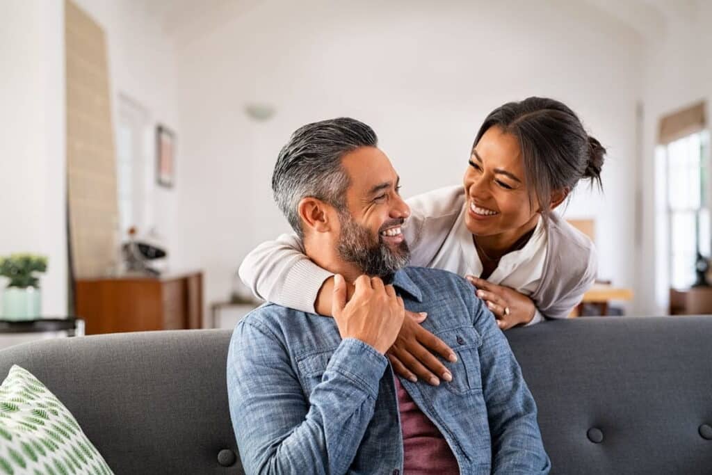 A shot of a happy couple looking at one another; the man is sitting on the couch, and the woman is hugging him from behind while he holds her arm.