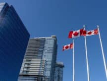 A shot of several skyscrapers in Vancouver, with three Canadian flags waving in the wind.