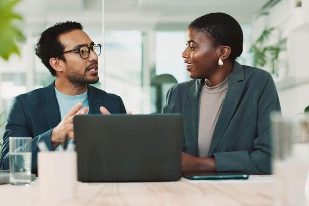 Man and woman having a discussion in an office environment, with a laptop and glass of water in front of them.
